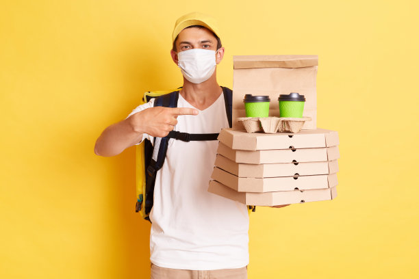 照片上的快递员背着保温背包，穿着白色t恤，戴着帽子和防护口罩，靠在黄色的墙上，指着披萨盒和装着咖啡的一次性杯子。图片下载