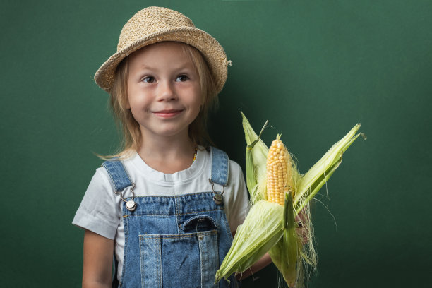 可爱的白人小女孩微笑着拿着玉米穗在深绿色的背景。快乐蹒跚学步的女孩戴着草帽和牛仔工装裤。全国玉米棒日快乐图片下载
