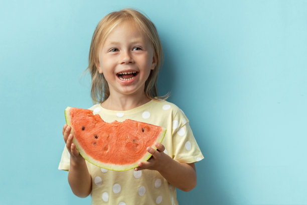 可爱的小女孩在蓝色的背景下享受着一大片鲜红多汁的西瓜。穿着黄色t恤的金发快乐白人女孩的肖像。炎热的夏天的概念图片下载
