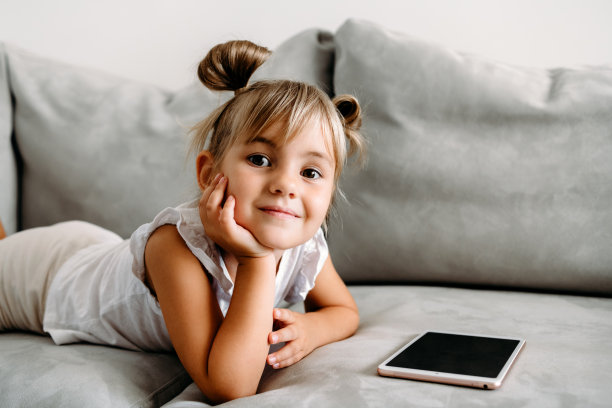 刚学步的小女孩躺在家里的沙发上玩数字无线平板电脑。宝宝的孩子成长与在线应用程序。儿童和电子设备的概念。婴儿肖像与智能手机。教育学习理念。图片下载