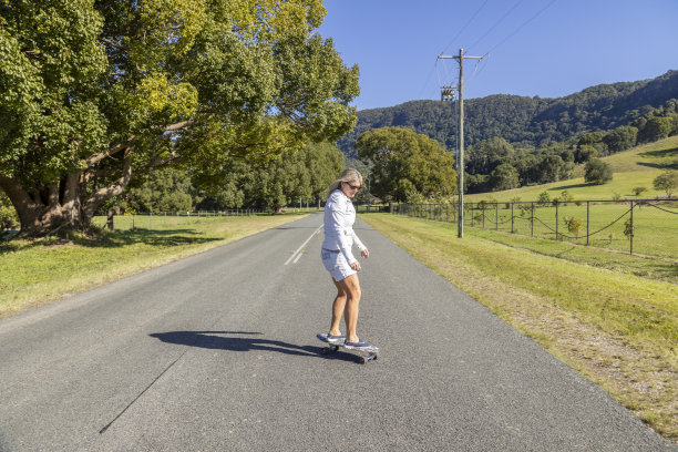 成熟的51岁金发女性骑着滑板在乡村道路上。牛仔短裤和白色夹克。一个金发女人在路中间玩滑板。澳大利亚昆士兰黄金海岸图片下载