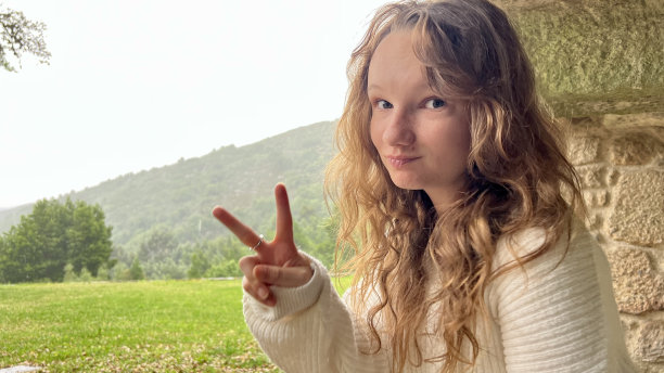 身穿白色夹克的金发少女站在山脉背景前，对着镜头皱着脸图片下载