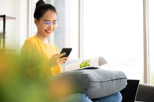 快乐的亚洲年轻女性在沙发上看书，在家里的客厅里拿着手机图片下载