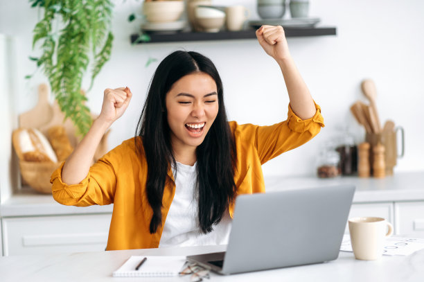 成功快乐情感的中国女孩，穿着时髦的衣服，自由职业者，IT专家，坐在厨房的桌子旁，拿着笔记本电脑，享受大利润，胜利，好消息，用拳头打手势，微笑图片下载
