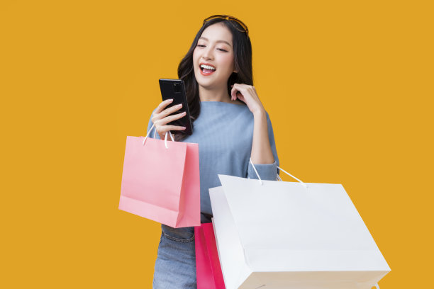 亚洲快乐女性女性女孩持有五颜六色的购物包站在黄色的背景工作室拍摄，特写肖像年轻漂亮有吸引力的女孩使用智能手机检查最好的交易销售图片下载
