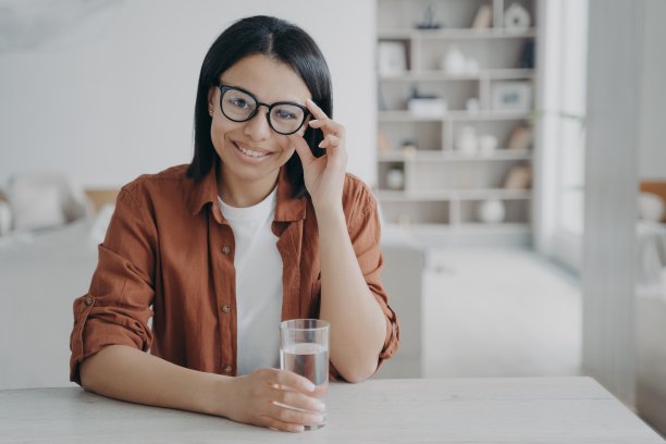 女孩喝水。年轻微笑的欧洲女人的肖像在眼镜，远程工作者在家里。图片下载