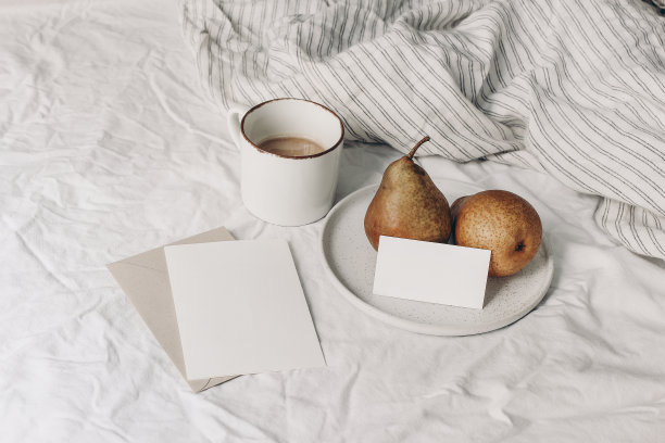 秋天的早餐在床上构成。一杯咖啡，空白的问候和名片模型。陶瓷盘子上的梨。白色条纹亚麻床上用品。生活方式的组合。内部。前视图。图片下载