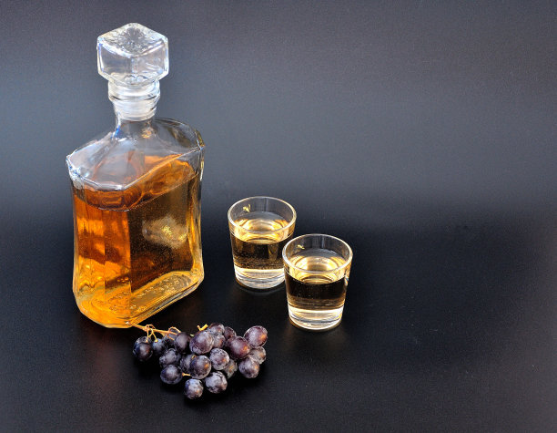 格拉帕酒(Grappa)，一瓶烈性酒精和两个玻璃杯放在黑色背景上，旁边放着一串黑色葡萄。图片下载
