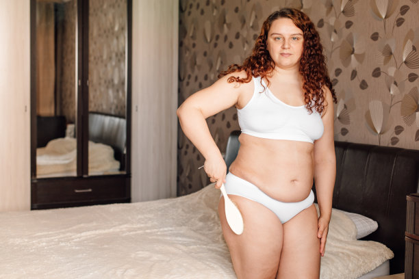 穿着白色内衣的卷发女士站在卧室的床边，用身体刷按摩臀部。大腿和腿粗的脂肪燃烧治疗。减肥程序，反脂肪程序图片下载