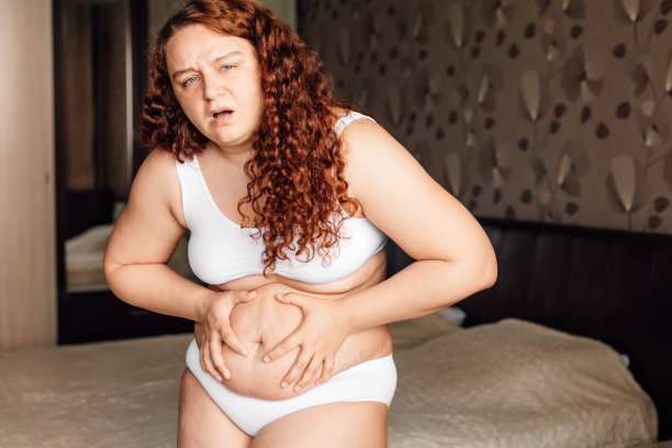 卷曲弯曲的大码女人穿着白色内衣站在卧室的床附近，忍受疼痛和抓在胃部特写。超重的女孩与肥胖作斗争。减肥计划健康问题图片下载
