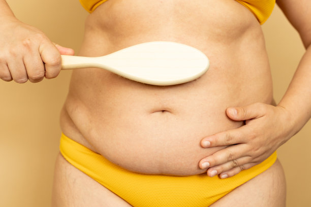 女人在黄色泳装按摩厚下垂的胃与身体刷特写，米色背景。减肥，对抗超重，肥胖和脂肪团。加上人的体型和身体的积极概念图片下载