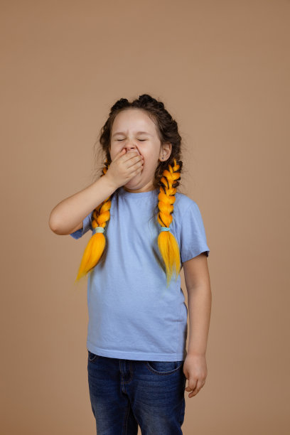 年轻疲惫疲惫的白人女孩与黄色的卡尼卡龙辫子打哈欠，闭上张开的嘴，闭上的眼睛穿着浅蓝色t恤上的米色背景图片下载