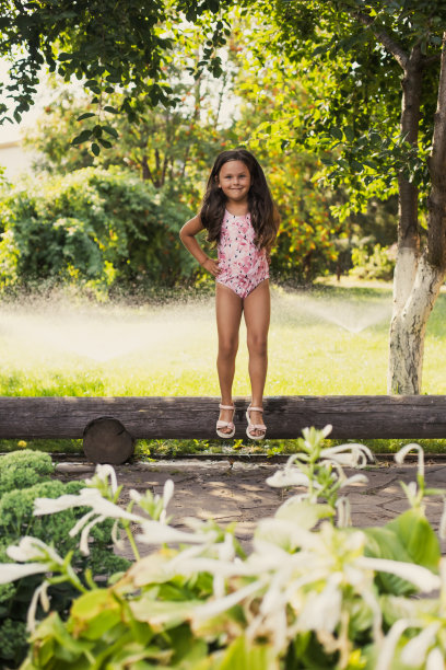 白天，以绿树和洒水器为背景的白人快乐女孩面带微笑地看着镜头，从圆木上跳下来。梦想成为体操运动员图片下载