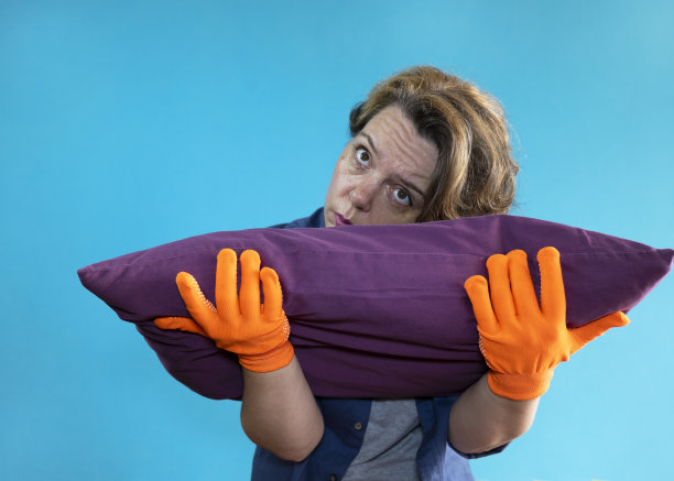 疲惫的女人戴着橙色手套，抱着蓝色背景下的紫色枕头。女人的肖像图片下载