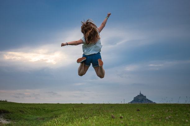 年轻的金发女孩纵身一跃，背景是圣米歇尔山。法国游客最多的地方之一图片下载