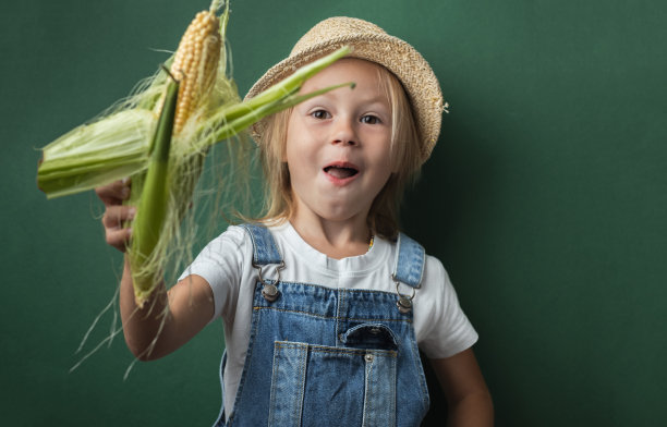 可爱的白人小女孩微笑着拿着玉米穗在深绿色的背景。快乐蹒跚学步的女孩戴着草帽和牛仔工装裤。全国玉米棒日快乐图片下载