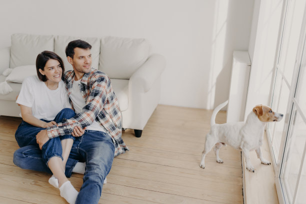 满意的浪漫情侣拥抱着看向窗外，在沙发旁的空房间里摆姿势，他们的狗想要去户外散步。女人和男人在家里享受业余时间，住在新的公寓图片下载
