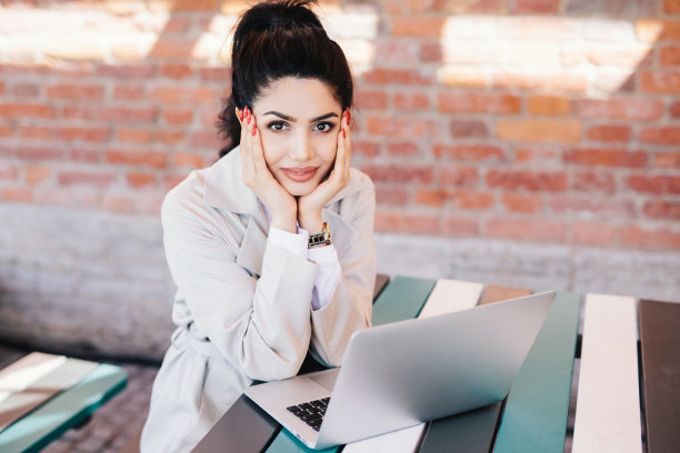 可爱的年轻黑发女子迷人的脸和红色的美甲穿着白色大衣坐在桌子上使用笔记本电脑聊天与她的家人使用免费的高速互联网连接图片下载