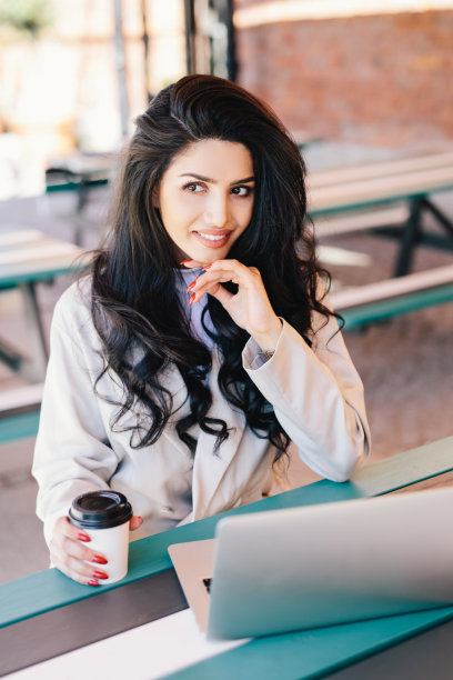 白种年轻女性自由职业者，波浪长发，黑眼睛，健康的皮肤，看起来很神秘，在现代笔记本电脑上远程工作，使用免费的无线网络喝着美味的咖啡。图片下载