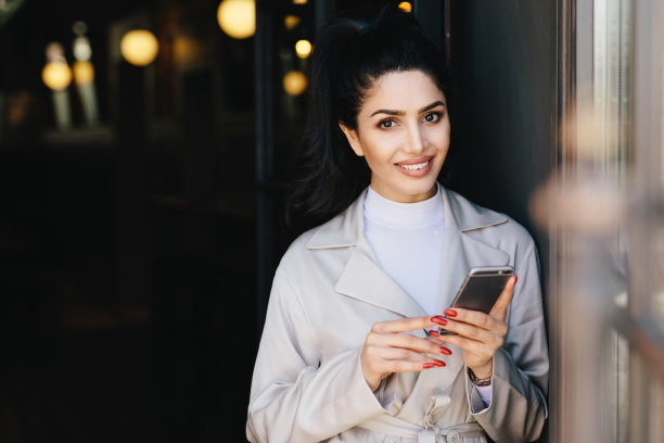 性感的黑发女士，迷人的黑眼睛和丰满的嘴唇，红色的指甲拿着手机，与她的朋友交流，微笑，看着相机。时尚的概念图片下载