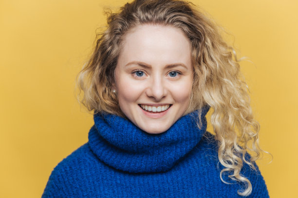 幸福快乐的女人，头发淡而挺拔，穿着温暖的蓝色毛衣，在成功的日子后有着宽阔的微笑和白色的牙齿，孤立在黄色的背景下。积极的情绪图片下载