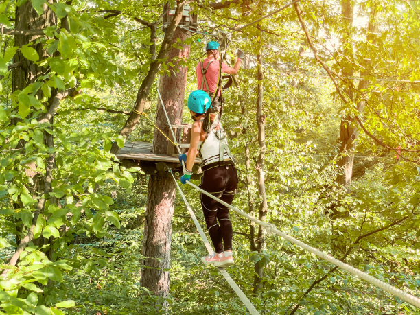 夏天的乐趣和体育冒险的人。十几岁的女孩在夏季森林的极限绳索公园的铰链小径。儿童在冒险跑道进行高空攀爬训练，配备安全带和防护头盔。图片下载