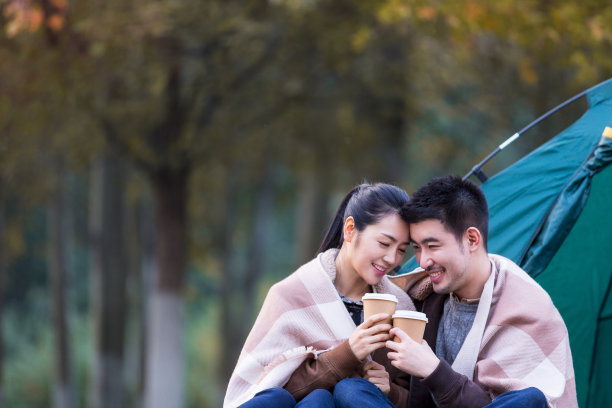 秋日夕阳下，一对年轻夫妇坐在彩色丛林草地上的露营帐篷前，围着同样的披肩喝咖啡——照片图片下载