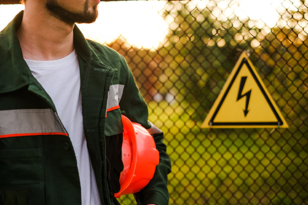 一名身穿制服、头戴安全帽的工程师站在电厂入口处，栅栏上有一个危险标志图片下载