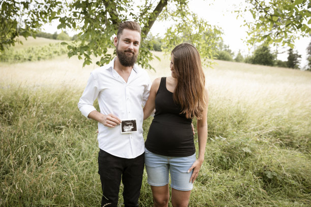 孕妇和她的丈夫站在绿色的草地上，抱着她宝宝的超声波照片图片下载