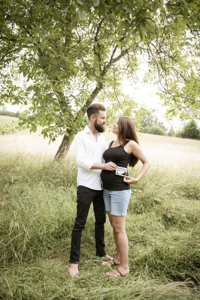 孕妇和她的丈夫站在绿色的草地上，抱着她宝宝的超声波照片图片下载