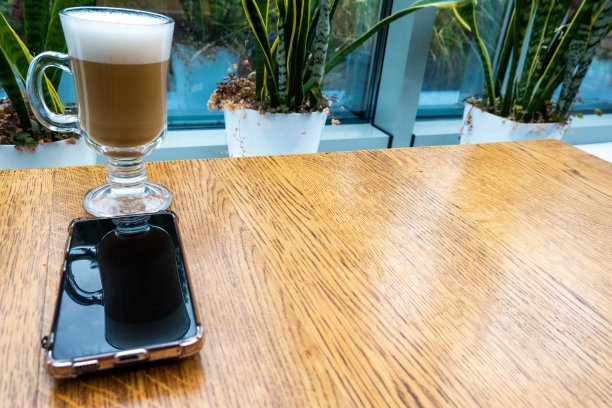 拿铁，泡沫在透明玻璃在餐厅的木桌上。室内有植物。智能手机黑屏上可见玻璃的剪影，躺在桌子上。镜像效应。舒适放松的餐厅氛围。图片下载