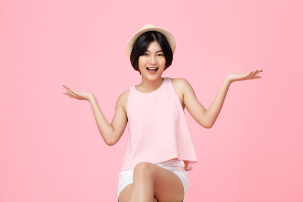 年轻漂亮微笑的亚洲女人坐在与开放的手势在粉红色的工作室孤立的背景图片下载