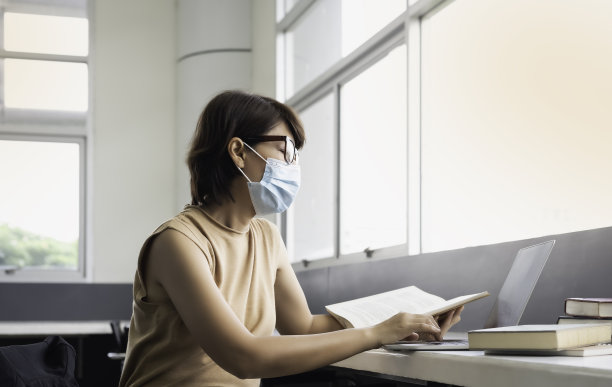 戴着眼镜和口罩的女人，在图书馆窗口边用笔记本电脑搜索资料图片下载