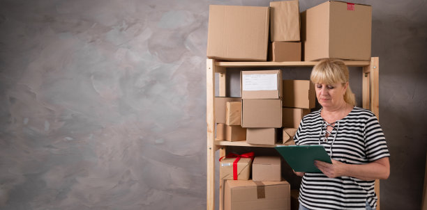 年长的女库存管理员在写字板上书写信息，成熟的女库存管理员在做仓库工作。企业主在家工作，在后台库存的包裹与产品准备出货。图片下载