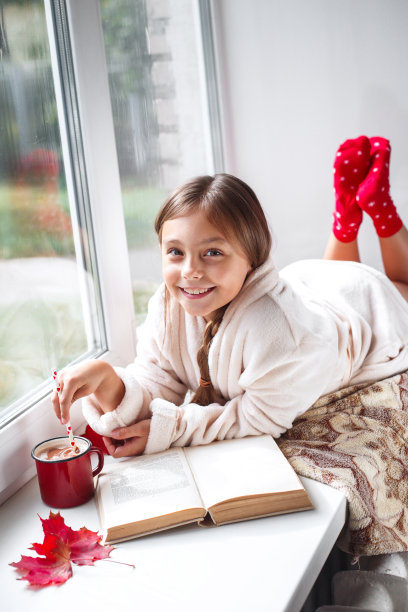 可爱的小女孩躺在窗户上，喝着可可，在家里的房间里看书。图片下载