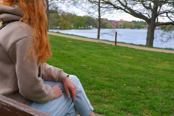 红头发的女人坐在湖边公园的长椅上。裁剪照片。图片下载