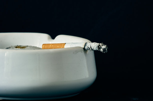 一根点燃的香烟躺在一个白色的陶瓷烟灰缸里，香烟慢慢地闷燃着，一长串烟灰已经出现图片下载