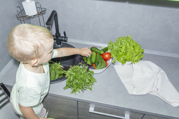 一个留着短发的小男孩在厨房帮忙做饭。在灰色的厨房里，在黑色的水槽里清洗新鲜的蔬菜和香草。图片下载