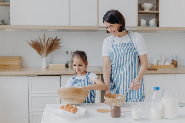照片中，深色头发的妈妈和孩子一起做曲奇饼，在碗里搅拌食材，穿着围裙，妈妈很高兴厨房里有个小帮手。一起烹饪，家庭和家庭的概念图片下载