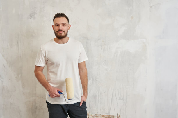 一个年轻人拿着一个涂色滚筒，背景是一堵白色的墙。维修、施工、刷墙图片下载