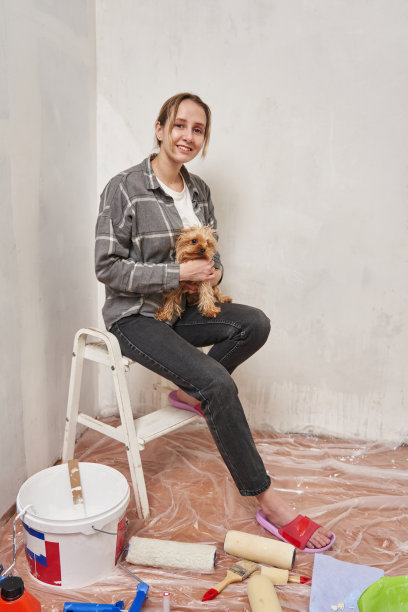 一个年轻的女人坐在一堵未粉刷的墙上的梯子上，手里拿着一只小狗。公寓装修概念图片下载