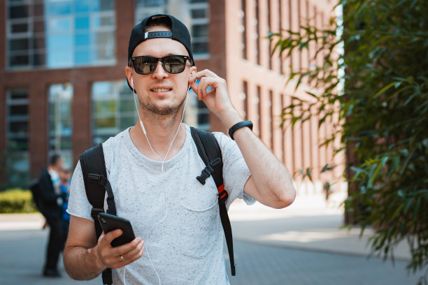 年轻人是一个时髦的家伙，戴着墨镜和黑色的帽子在城市里走，手里拿着智能手机，听着音乐耳机。T图片下载
