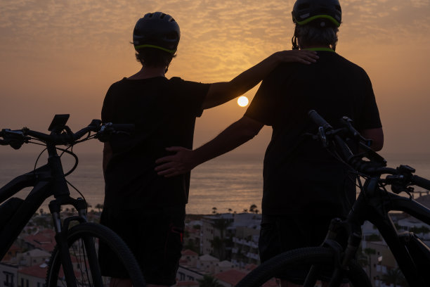 夕阳下，戴着自行车头盔、骑着电动自行车站在山坡上的老年运动夫妇剪影，是退休人士享受假期的健康活动图片下载
