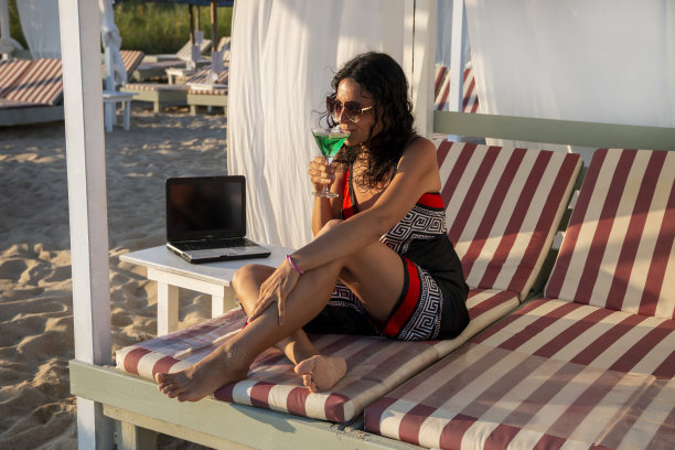商务女性用笔记本电脑躺在沙发床上，在海滩上喝鸡尾酒图片下载