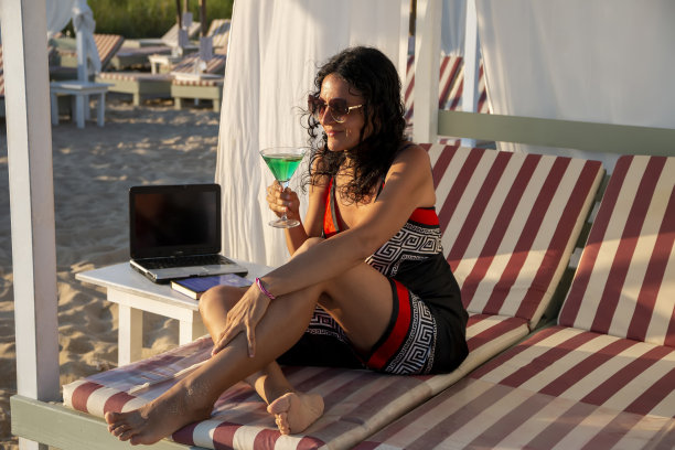 商务女性用笔记本电脑躺在沙发床上，在海滩上喝鸡尾酒图片下载