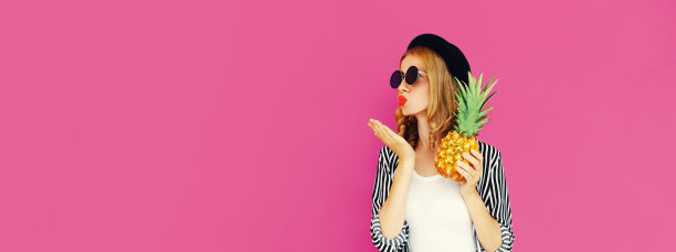 夏天的肖像美丽的年轻女子菠萝吹她的嘴唇发送甜蜜的空气吻戴着黑色的帽子在粉红色的背景，空白的拷贝空间的广告文字图片下载