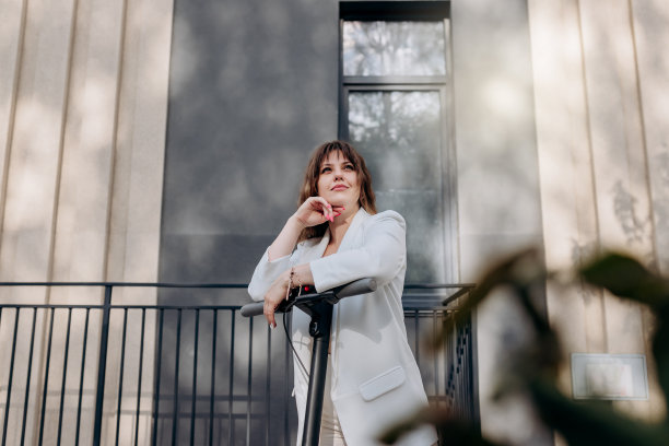 美丽的年轻女子穿着白色西装站在她的电动机车附近的现代建筑在城市和望向别处图片下载