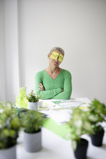 短发的中年妇女坐在摆满绿色植物和工作笔记的办公桌前，脸上画着眼睛的便签纸图片下载