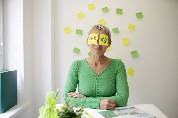 短发的中年妇女坐在摆满绿色植物和工作笔记的办公桌前，脸上画着眼睛的便签纸图片下载