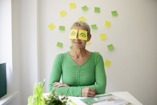 短发的中年妇女坐在摆满绿色植物和工作笔记的办公桌前，脸上画着眼睛的便签纸图片下载